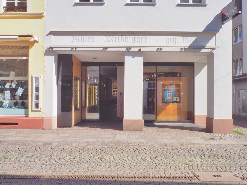 Ein weißes Ladengeschäft mit großen Fenstern, Glastür und der Aufschrift "Brillen Weissburger gegr. 1878" über dem Eingang