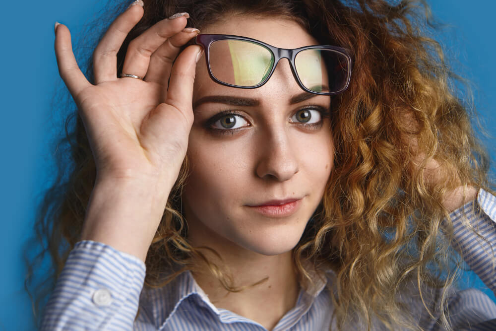 Eine Frau mit lockigen Haaren hebt ihre Sonnenbrille und schaut nach vorn, trägt ein gestreiftes Shirt vor blauem Hintergrund zum Thema Winkelfehlsichtigkeit