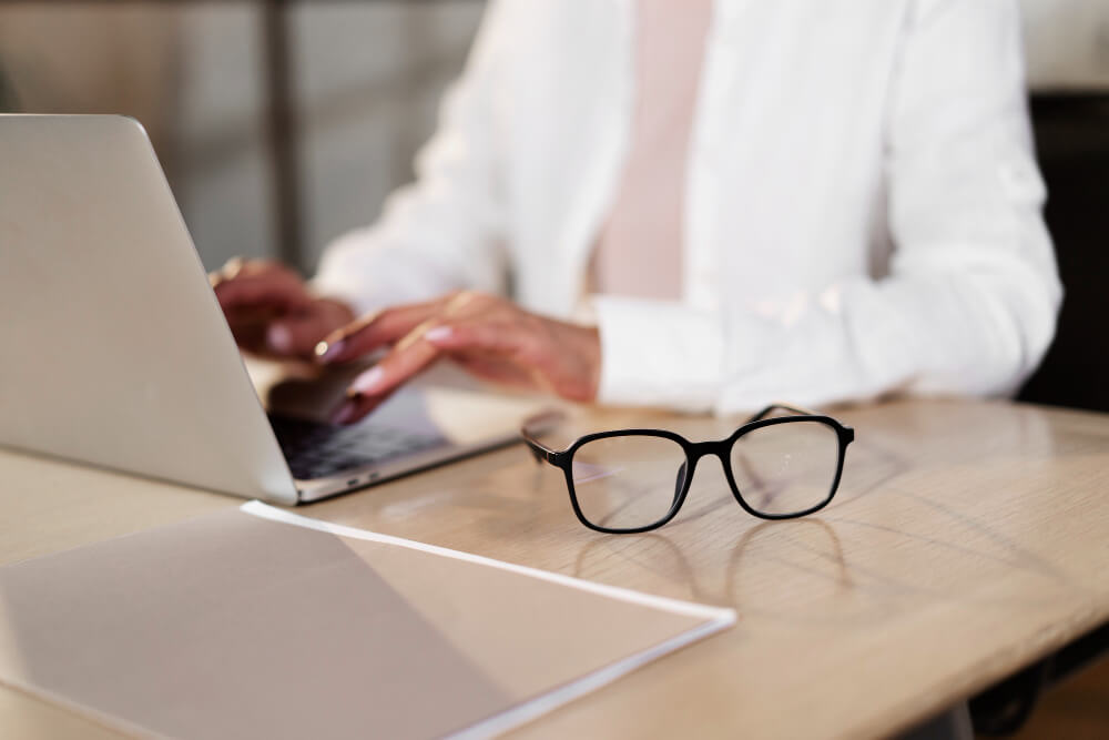 An einer Schreibtisch-Arbeitsplatzsituation tippt jemand am Laptop, eine Arbeitsplatzbrille liegt griffbereit daneben