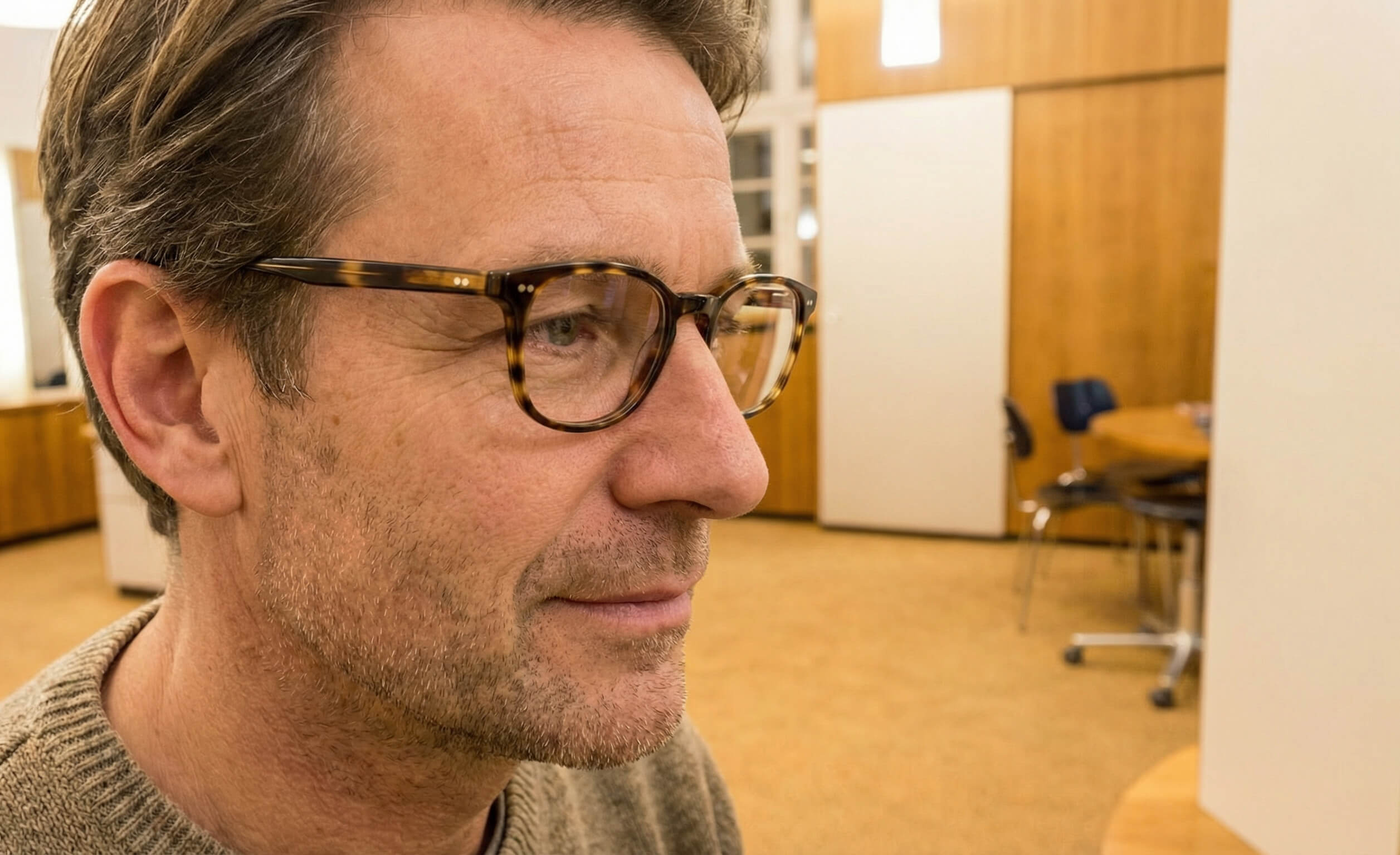 Ein Mann mit Brille und Bartstoppeln trägt einen Pullover und wirkt nachdenklich in einem warm beleuchteten, holzgetäfelten Büro