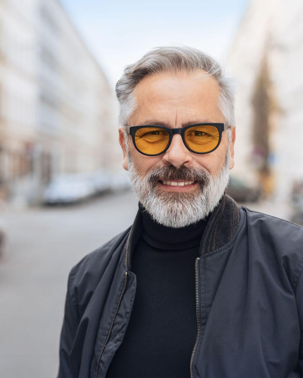 Ein lächelnder Mann mit grauen Haaren, Bart und gelber Brille trägt eine schwarze Jacke und steht auf einer belebten Stadtstraße