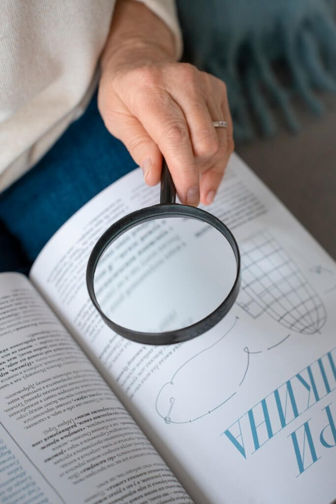 Eine Person hält eine Lupe über ein aufgeschlagenes Buch, wodurch Text und eine Abbildung auf der Seite vergrößert werden