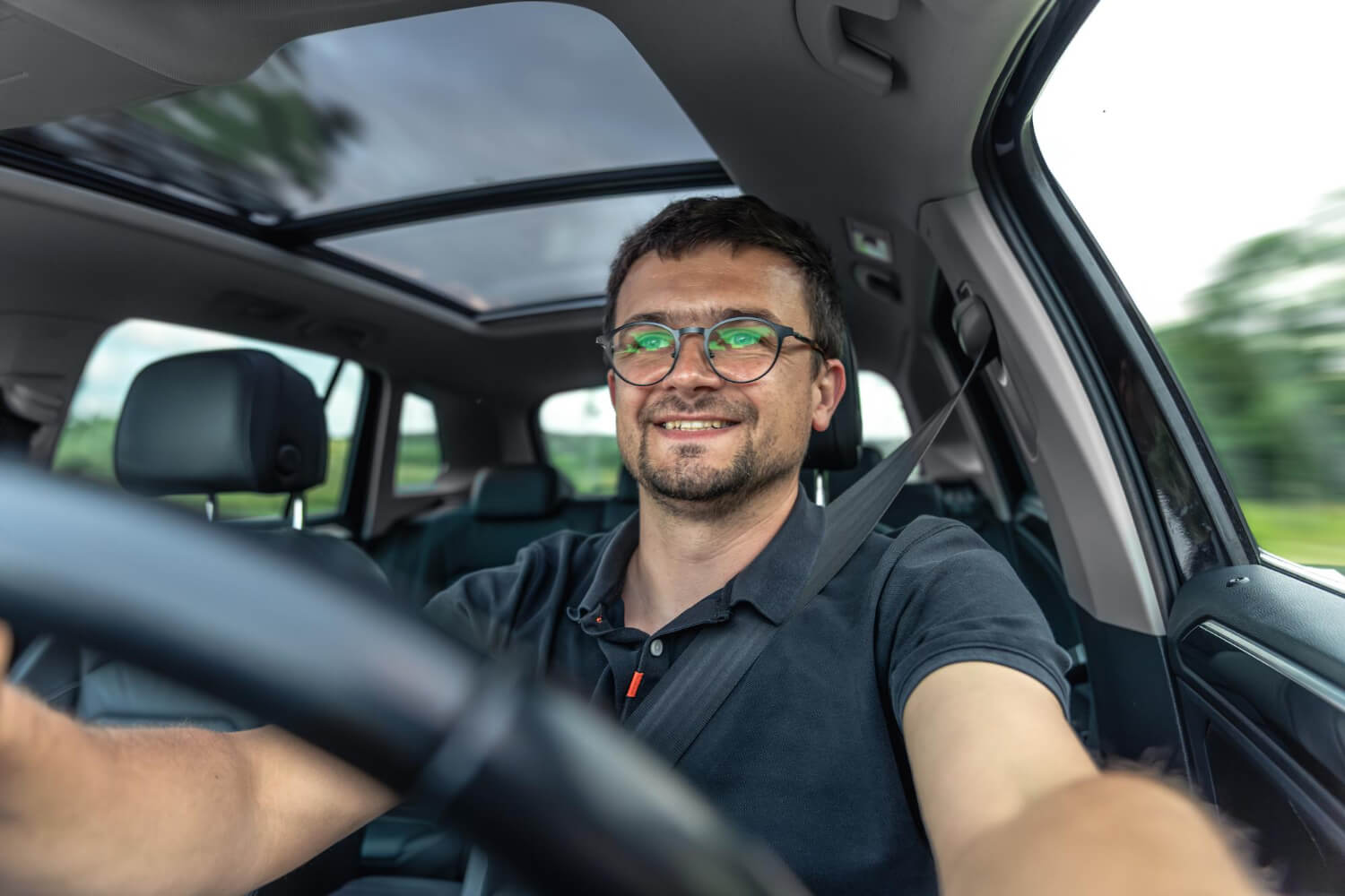 Ein Mann mit Gleitsichtbrille und angeschnallt lächelt beim Fahren eines Autos mit Schiebedach an einem sonnigen Tag