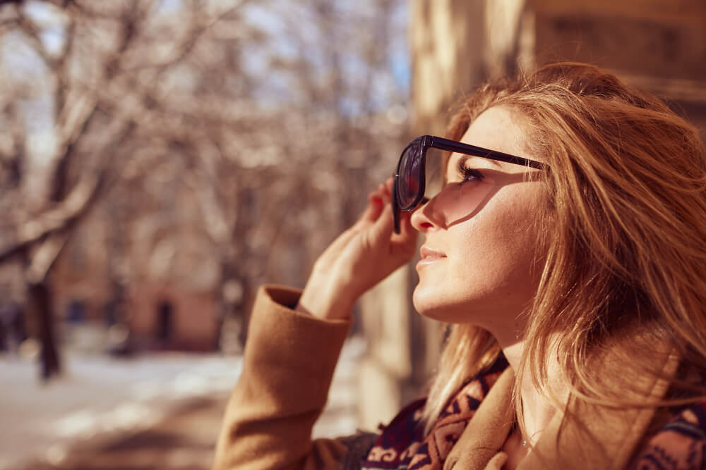 Eine Frau im Mantel rückt an einem sonnigen, verschneiten Tag ihre Sonnenbrille zurecht - ein automatisch gespeicherter Entwurf für Wintermode