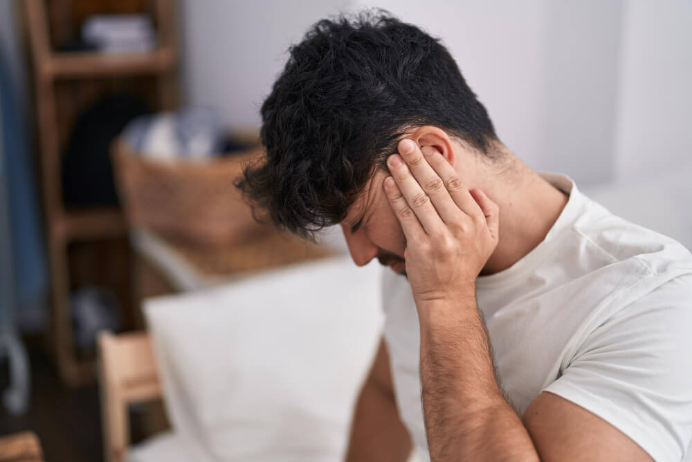 Ein Mann sitzt auf einem Bett, hält sich schmerzerfüllt das Ohr und blickt nach unten, automatisch gespeicherter Entwurf