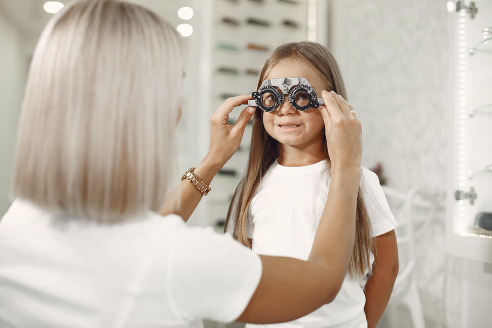 Eine Frau hilft einem Mädchen beim Aufsetzen eines Sehtestgeräts, das Kind braucht eine Brille beim Sehtest im Optikerbüro