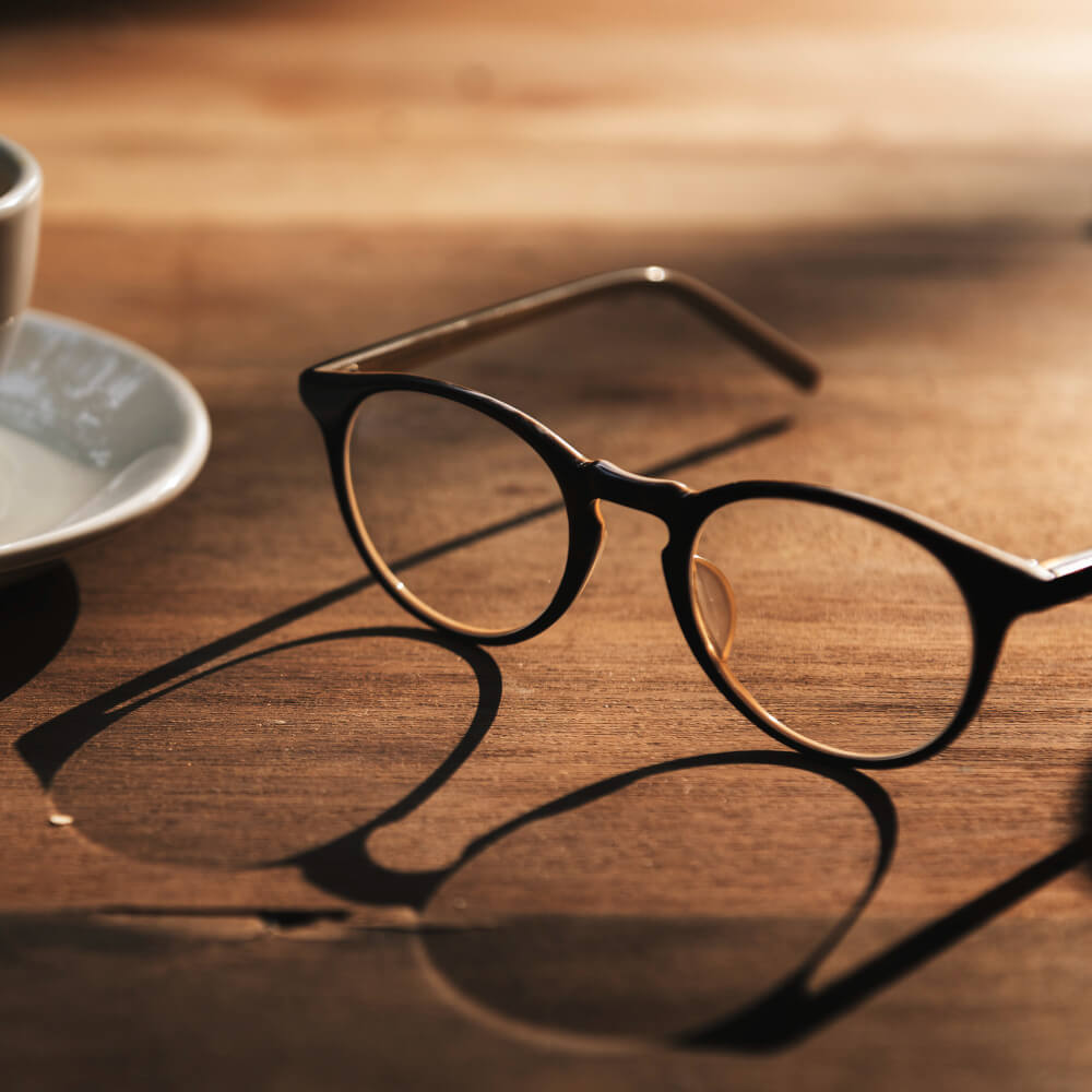 Ein rundes Brillenpaar liegt auf einem sonnenbeschienenen Holztisch neben einer weißen Untertasse