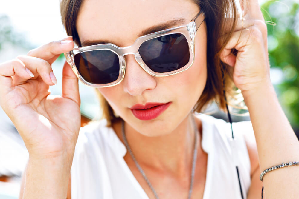 Eine Frau im weißen Top richtet ihre Sonnenbrille mit Sehstärke und blickt draußen im sonnigen Karlsruhe in die Kamera