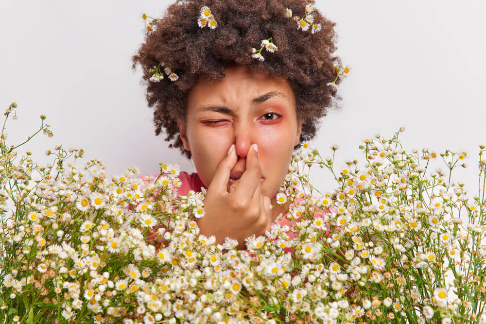Eine Person mit Blumen im Haar hält sich hinter Gänseblümchen die Nase zu, eventuell wegen einer Pollenallergie oder Kontaktlinsen