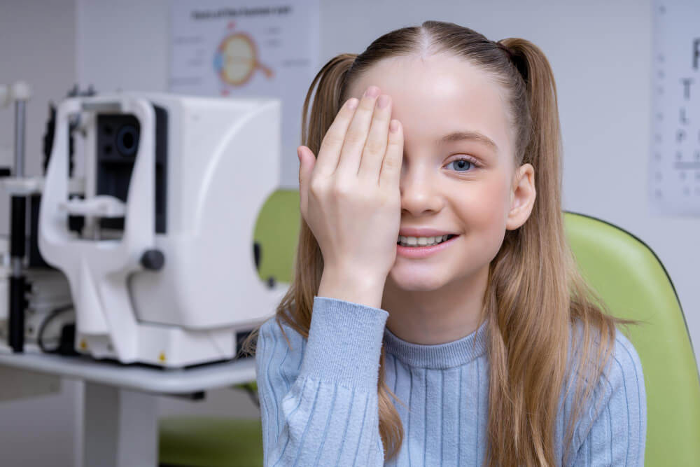 Ein blondes Mädchen hält sich bei einer Augenuntersuchung im Optikstudio ein Auge zu und lächelt, automatisch gespeicherter Entwurf