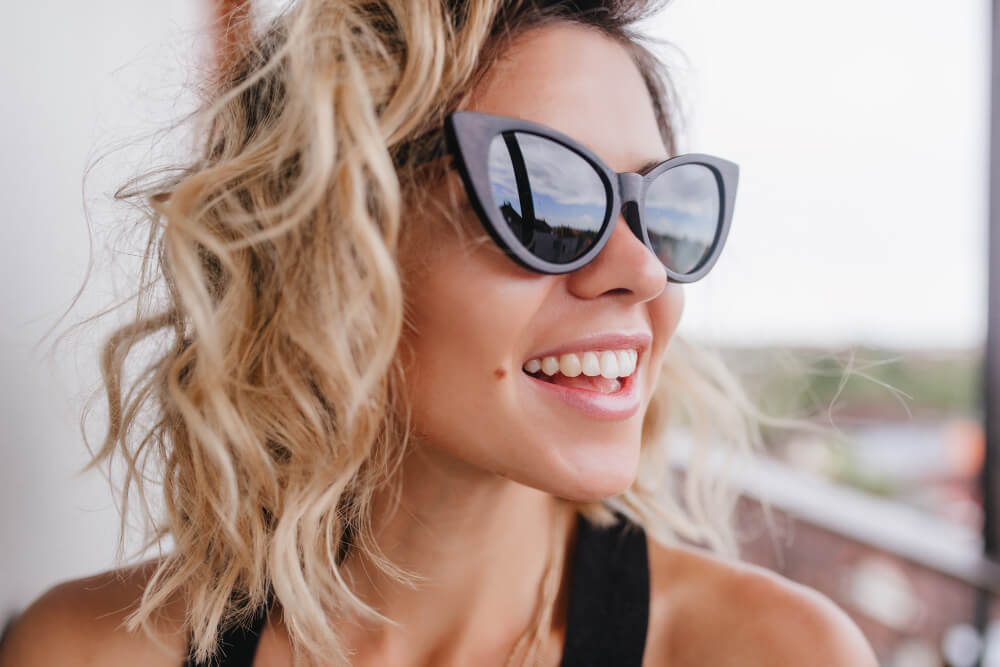 Draußen steht eine lächelnde Frau mit welligen blonden Haaren und Cat-Eye-Sonnenbrille, sie trägt ein schwarzes Oberteil