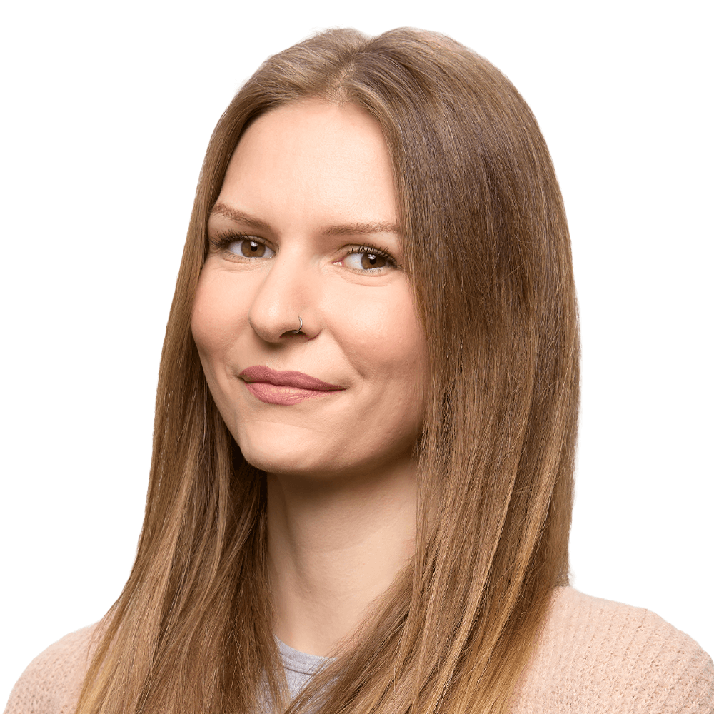 Eine Frau mit langen glatten braunen Haaren, Nasenring und hellem Pullover lächelt sanft vor weißem Hintergrund
