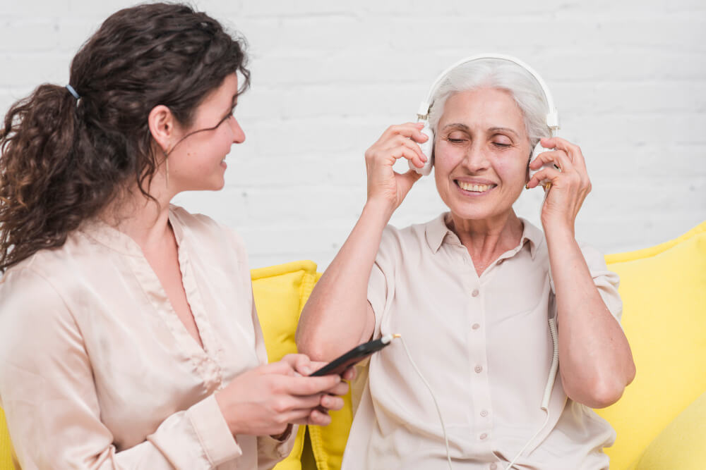 Zwei Frauen sitzen auf einem gelben Sofa; die eine genießt mit Kopfhörern die Musik, die andere stellt die Lautstärke an ihrem Smartphone ein