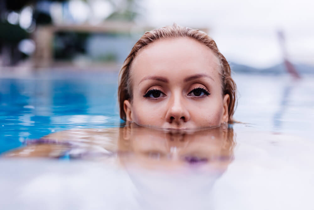 Eine Frau mit nassen Haaren taucht teilweise im Pool, nur Augen und Stirn sind über Wasser und sie schützt ihre Augen im Wasser